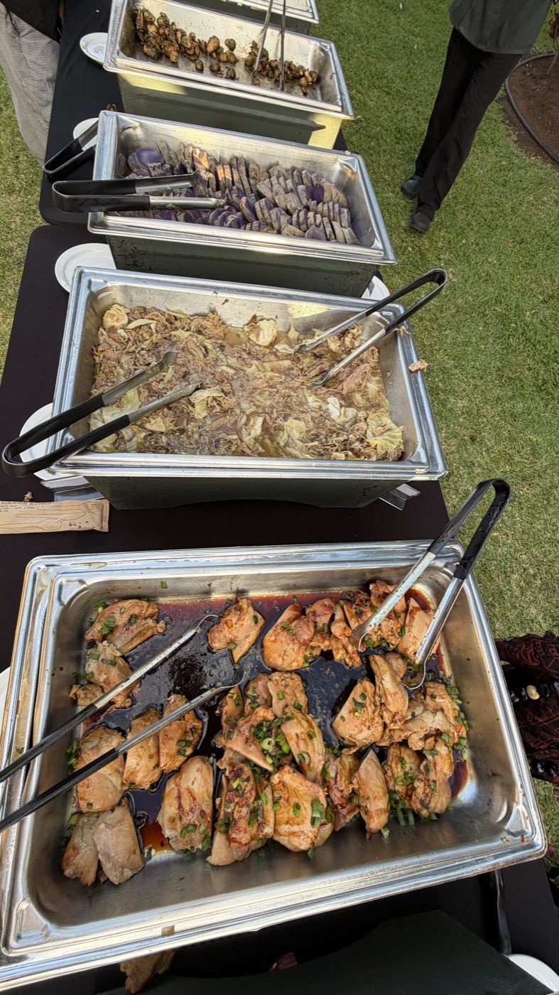 A row of open metal buffet trays with cooked food, serving tongs, and spoons
