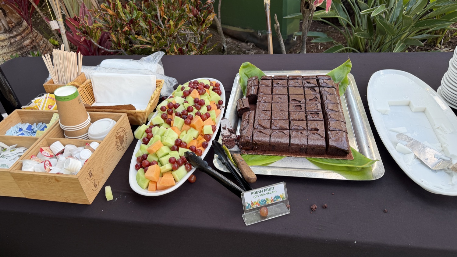 A dessert table with a platter of cut fruit, a tray of chocolate squares, cups, and serving tongs