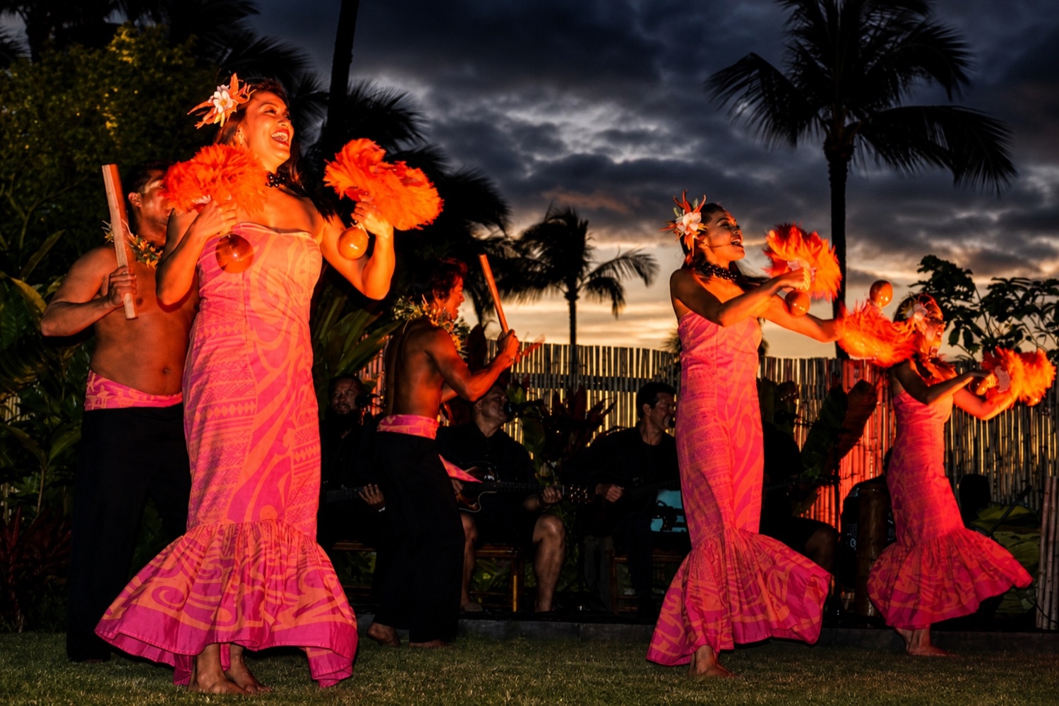 Dancers in pink dresses hold orange feathered rattles beneath palm trees