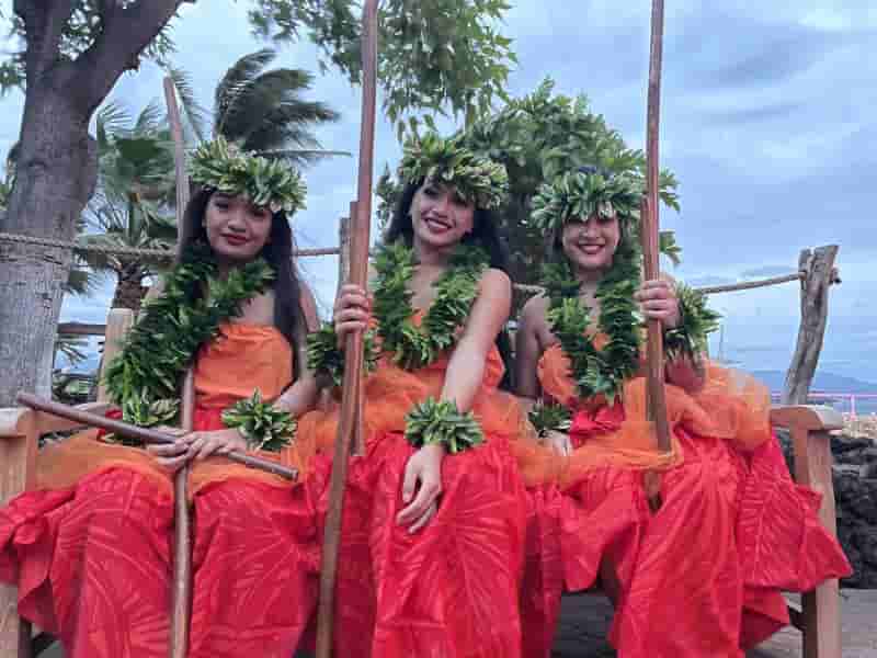 Three hula dancers seated in traditional orange dress with green lei headpieces