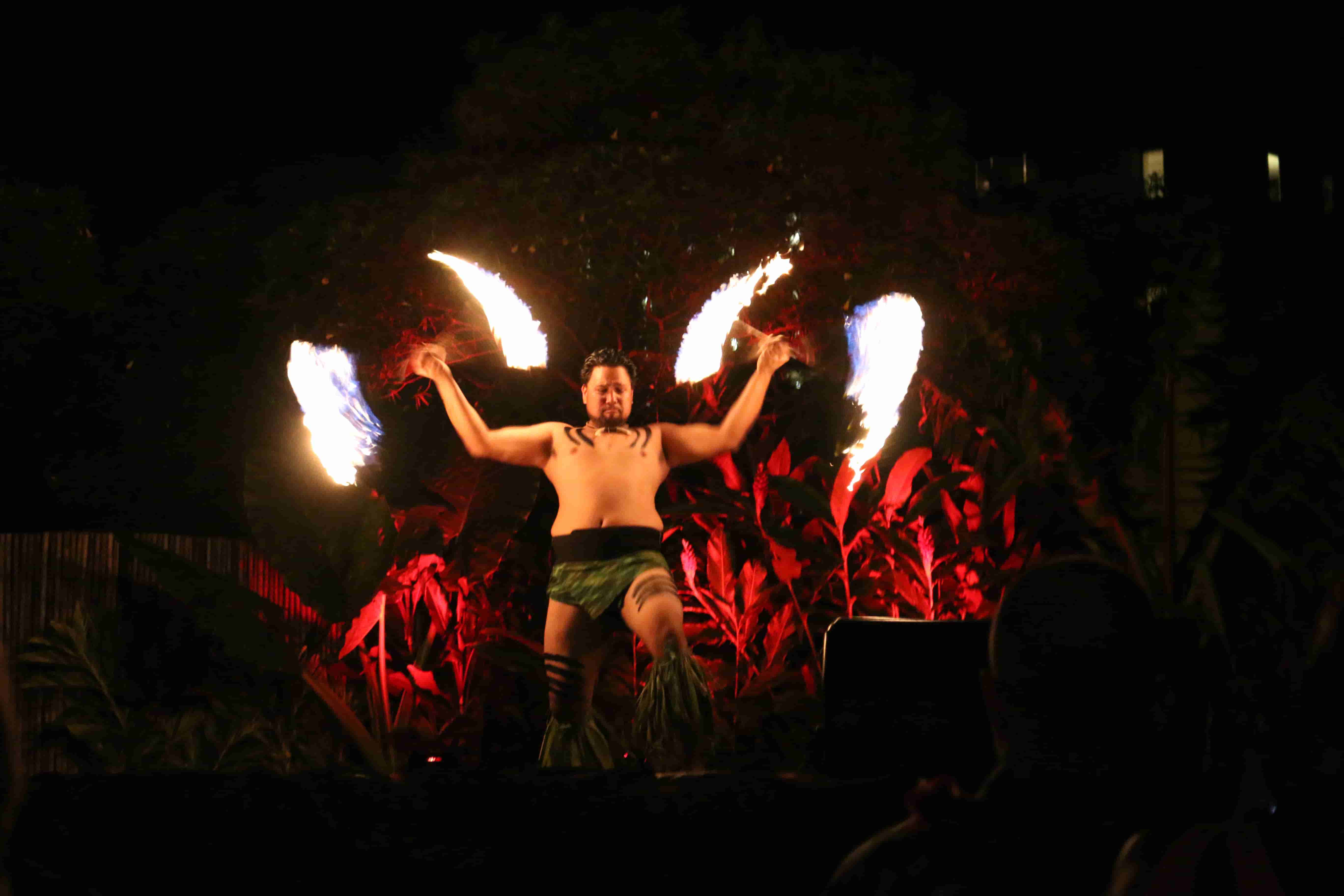 Fire performer with dual flaming knives against red-lit foliage